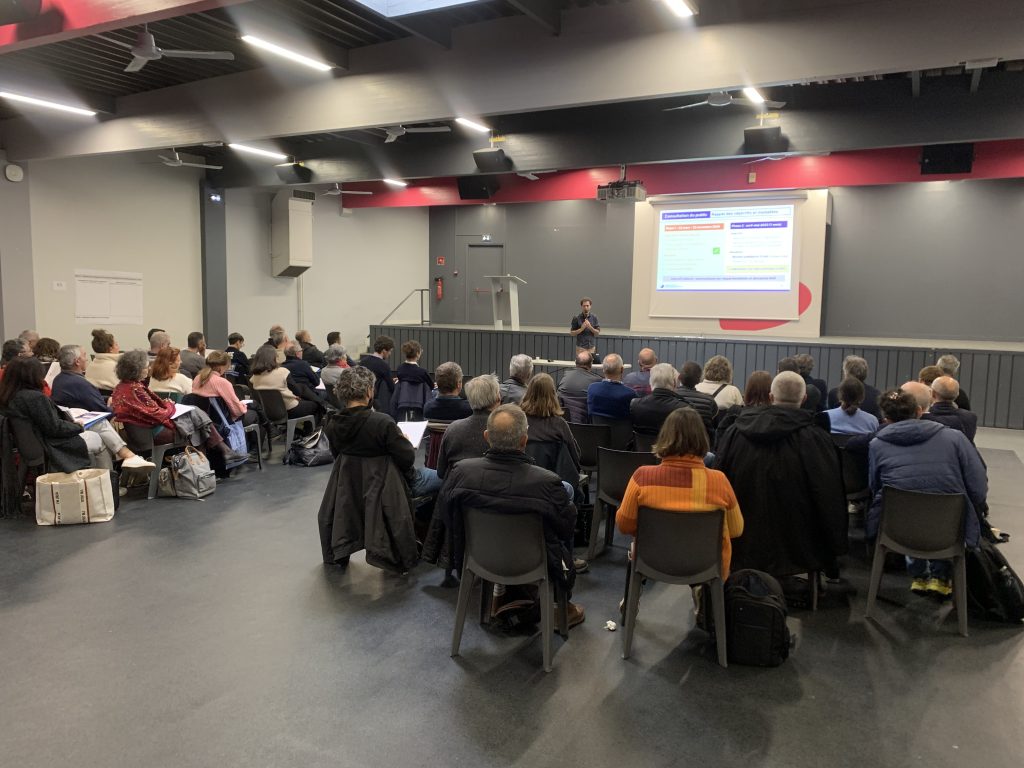 Photo d'une réunion de consultation organisée dans la cadre de l'élaboration du PAPI complet. Les citoyens, en plénière, écoutent l'animateur, face à un écran diffusant un support de présentation.
